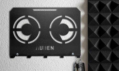 Metalowy wieszak na klucze z geometrycznym wycięciem i spersonalizowaną nazwą, zamontowany na teksturowanej ścianie