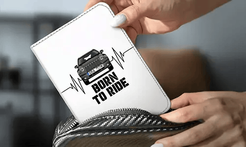 Persona sostiene una cartera blanca con coche negro y diseño de latido, texto BORN TO RIDE, fondo con funda para permiso con matrícula y coche de fibra de carbono